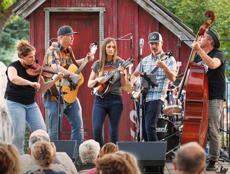 Bridging Betty performing outdoors at golden hour in front of a red barn: a five-piece Americana band with fiddle, guitar, mandolin, banjo, and upright bass, with audience visible in the foreground.