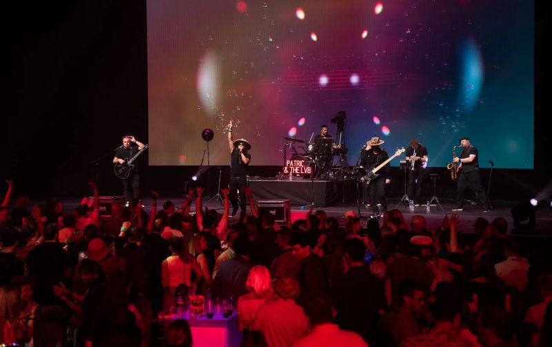 Patrick & the LVB on stage at Dolby Live in Las Vegas, seven-piece band silhouetted against a massive LED screen with a packed crowd of fans cheering in the foreground.
