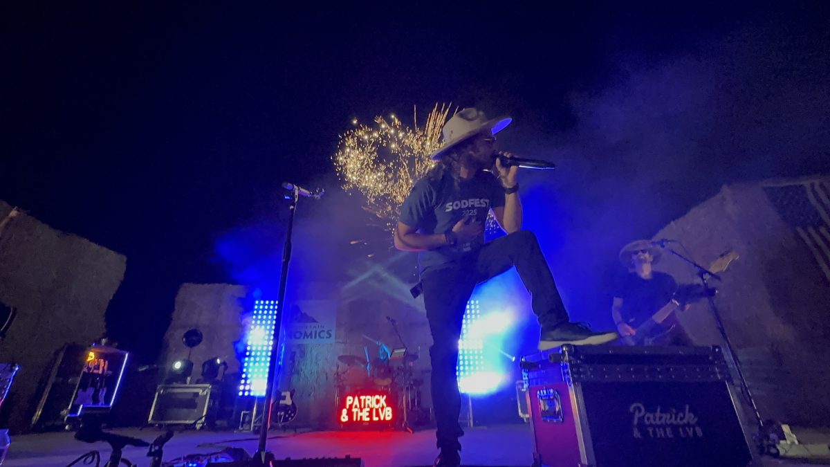 Patrick stands one foot up on a road case in a SODFEST 2025 t-shirt and cowboy hat, mic to his mouth, white fireworks bursting in the night sky behind the hay-bale stage.